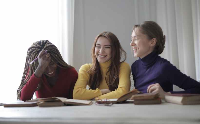 cheerful young multiethnic women having fun while sitting with books at table in modern workspace